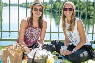 Zwei junge Frauen sitzen auf einer Picknickdecke, im Hintergrund ist ein Fluss zu sehen